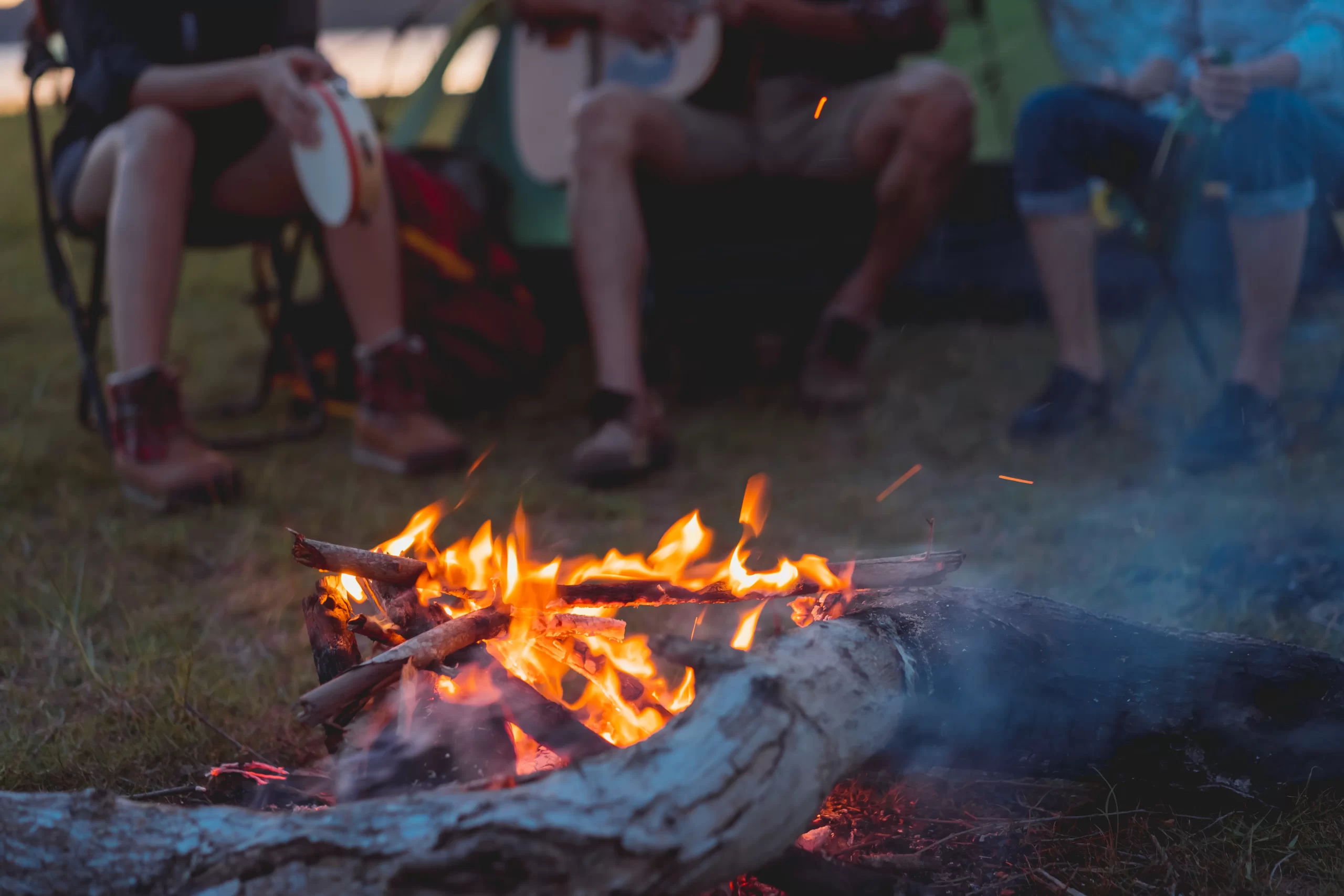 A group of people sits around a campfire near the bayview in the evening. Flames and sparks rise from the burning logs, while some play musical instruments like a tambourine and guitar. The scene suggests a relaxed, outdoor setting reminiscent of nights at a cozy cabin by Crosslake.