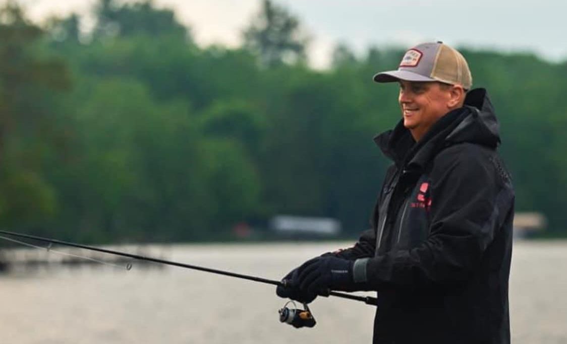 A person wearing a cap and jacket is fishing by a tranquil lake at Crosslake Resort. They are holding a fishing rod, with a blurred backdrop of trees and water creating a serene view.